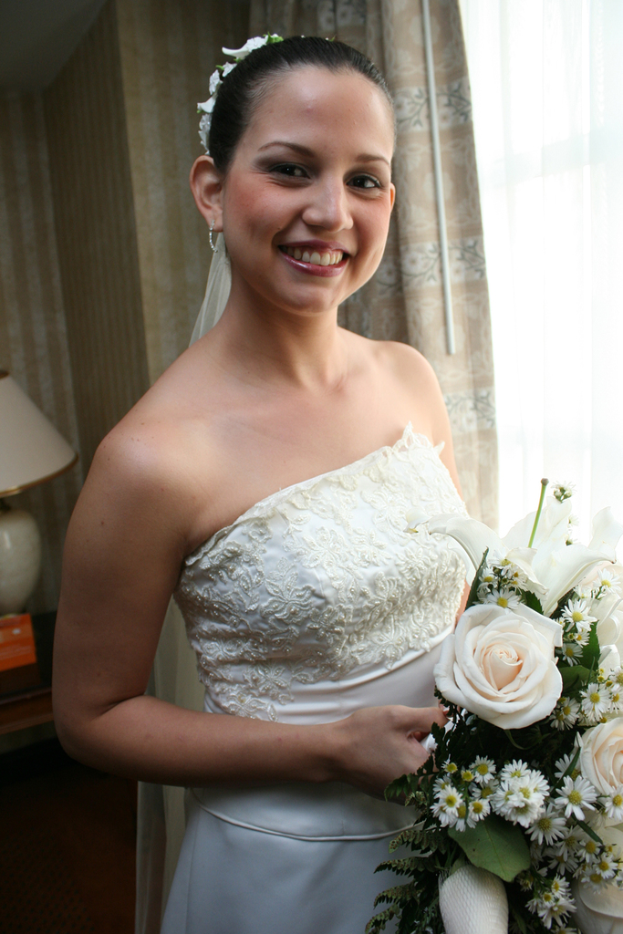Eine Braut steht strahlend in der Mitte, hält einen Strauß in der Hand, mit einem Vorhang und einer Wand im Hintergrund und einer Lampe auf einem Tisch links.