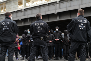 Eine Gruppe von Polizisten in Uniform steht vor einer Menge von Menschen, die ebenfalls schwarze Uniformen und Masken tragen, mit einer Brücke und einem Gebäude im Hintergrund, was auf eine städtische Umgebung hinweist.
