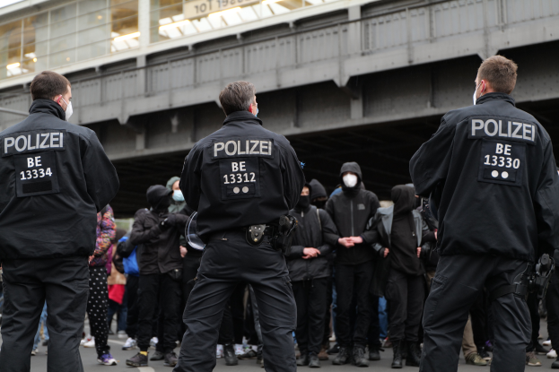 Eine Gruppe von Polizisten in Uniform steht vor einer Menge von Menschen, die ebenfalls schwarze Uniformen und Masken tragen, mit einer Brücke und einem Gebäude im Hintergrund, was auf eine städtische Umgebung hinweist.