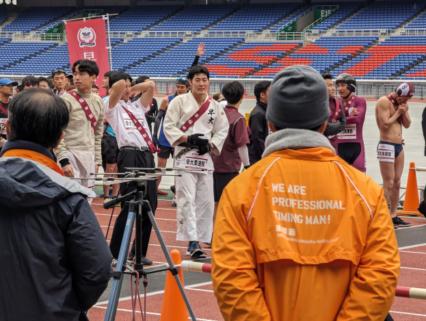 Eine Gruppe von Menschen auf einer Bahn mit einer Kamera auf einem Stativ im Vordergrund, Stühlen, einem Banner und Lichtern im Hintergrund.