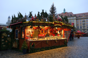 Ein Weihnachtsmarktstand auf einem Kopfsteinpflasterweg, umgeben von Gebäuden, geschmückten Bäumen und Menschen, unter einem sichtbaren Himmel.