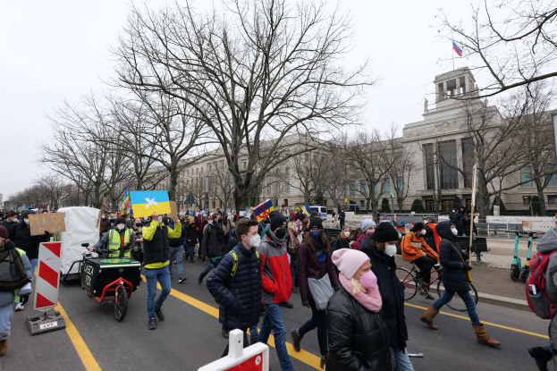 Eine große Gruppe von Menschen marschiert auf einer Straße in Washington, D.C., mit Schildern und Bannern, während einige Fahrräder fahren, unter einem klaren blauen Himmel.