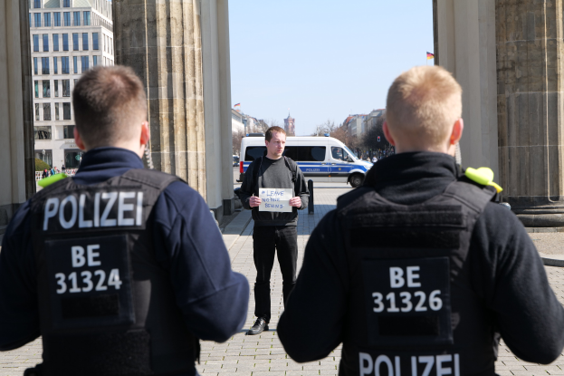 Drei Polizisten stehen vor einem Gebäude, wobei ein Mann in der Mitte ein Blatt Papier hält, flankiert von Säulen und umgeben von Fahrzeugen, Menschen, Bäumen und Gebäuden unter dem Himmel.