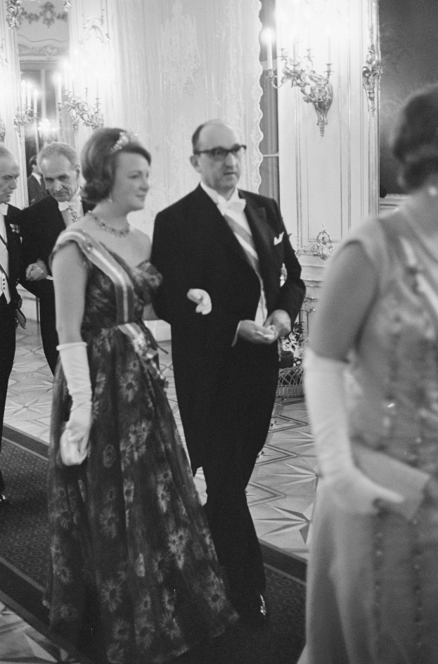 Ein Mann in einem schwarzen Blazer, weißem Hemd und schwarzen Hosen sowie eine Frau in einem eleganten Kleid gehen einen von Wandlampen beleuchteten Flur entlang, umgeben von einer Gruppe von Menschen, die auf dem Boden stehen.