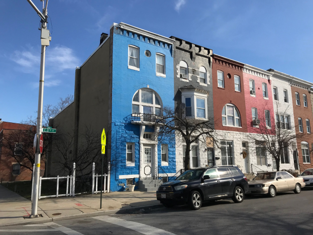 Eine Reihe bunter Häuser mit parkenden Autos auf einer Straße, gesäumt von Bäumen, Pfählen, Brettern und einem Zaun unter einem bewölkten Himmel.