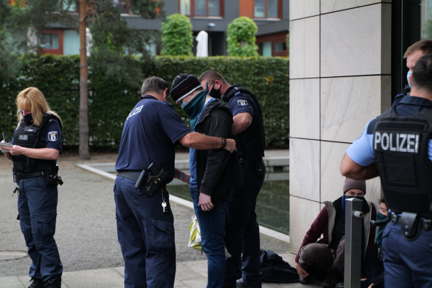 Gruppe von Polizeibeamten vor einem Gebäude stehend, mit einer Frau, die ein Buch und einen Stift hält, und mehreren Menschen in der Nähe; Hintergrund enthält Pflanzen, Bäume und Gebäude.