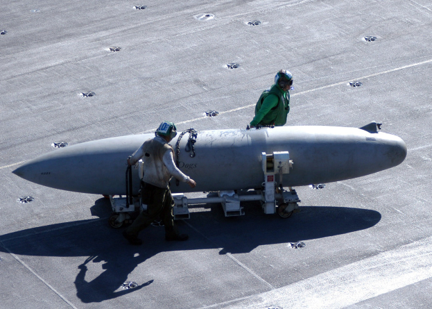 Zwei Männer in Schutzausrüstung inspizieren eine große, spitz zulaufende zylindrische Rakete, die einer Atombombe ähnelt, auf einem Flugzeugträger.