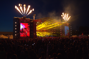 Eine große Menschenmenge auf einem Musikfestival mit einer hell erleuchteten Bühne, auf der Musiker spielen, sowie Zelte und Feuerwerk am dunklen Himmel.