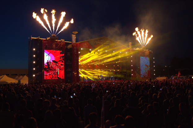 Eine große Menschenmenge auf einem Musikfestival mit einer hell erleuchteten Bühne, auf der Musiker spielen, sowie Zelte und Feuerwerk am dunklen Himmel.
