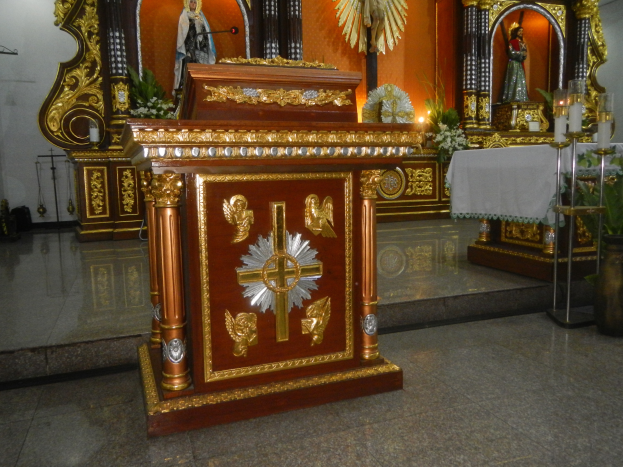 Ein Altar in einer Kirche mit einem Kreuz, Statuen, Blumenbouquets, Kerzen, einem tuchbedeckten Tisch und anderen Gegenständen, mit Wänden, die mit Lichtern geschmückt sind.