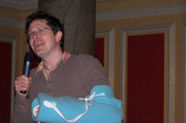 Ein Mann steht drinnen und h√§lt ein Mikrofon in seiner rechten Hand und einen Frakturfixiergurt in seiner linken, tr√§gt ein braunes T-Shirt und Brille, mit einer S√§ule und Wand dahinter.