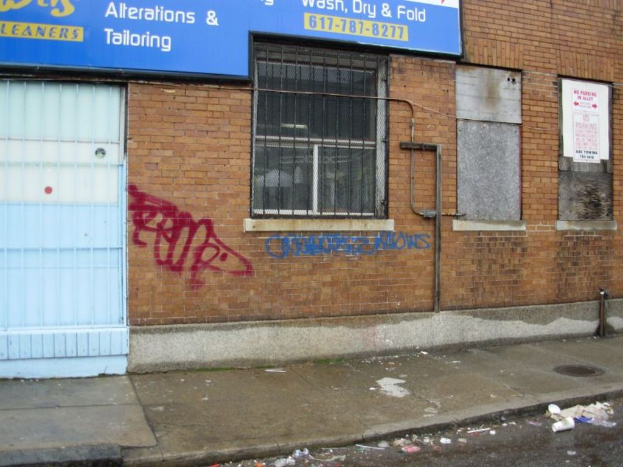 Eine Straßenszene mit einem Fußweg und einer Straße, die mit Müll übersät sind, einer Backsteinwand, Fenstern, Rohren, einem Tor und einer Werbetafel oben.