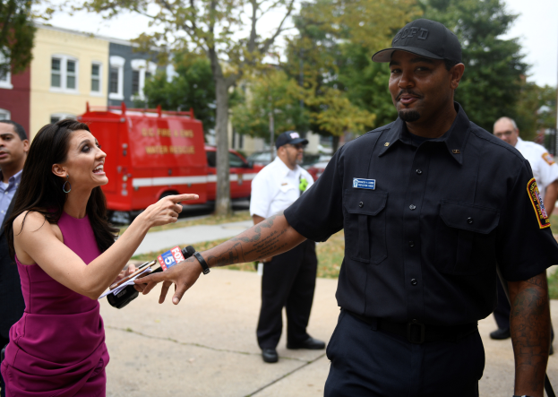 Eine Frau in einem lilafarbenen Kleid spricht mit einem Polizisten in einem schwarzen Kleid und einer Mütze, mit mehreren Personen, Fahrzeugen, Bäumen, Gebäuden und einem weißem Himmel im Hintergrund.
