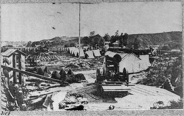 Schwarz-weiß-Foto einer Baustelle mit einer Maschine im Vordergrund, umgeben von Hütten, Bäumen, Hügeln und einem Himmel im Hintergrund, mit Text unten, der lautet: "Die Bauarbeiten an einer neuen Brücke in der Stadt San Francisco, Kalifornien."