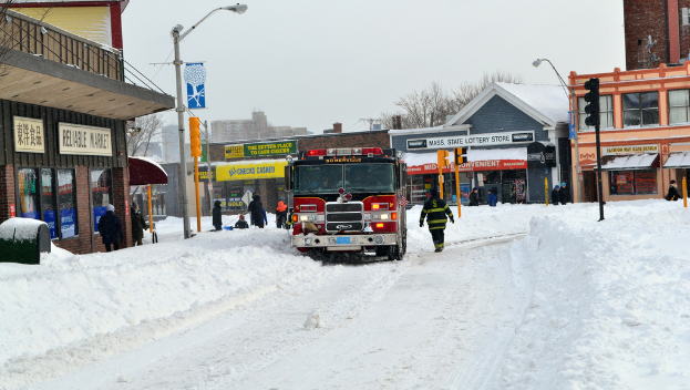 Eine Feuerwache steht im Schnee vor Gebäuden, mit Geschäften und Laternenmasten links, einer Ampel in der Mitte und Schnee auf dem Boden.