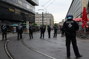 Gruppe von Polizeibeamten in schwarzen Uniformen und Helmen auf einer Straße mit Gebäuden, Glasfenstern und verstreuter Außenmöblierung unter einem klaren Himmel.