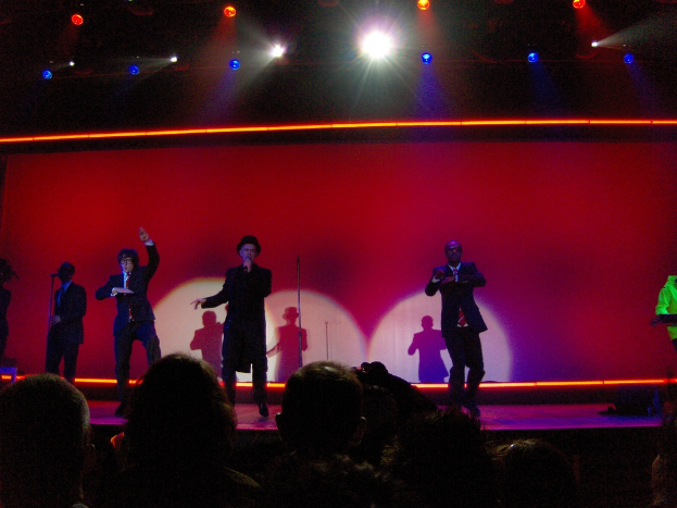 Ein Mann in einem schwarzen Kleid hält ein Mikrofon auf der Bühne, mit drei Tänzern neben ihm; ein Kameramann und Zuschauer vorne und ein Pianist in einem grünen Jackett rechts, mit Scheinwerfern oben.
