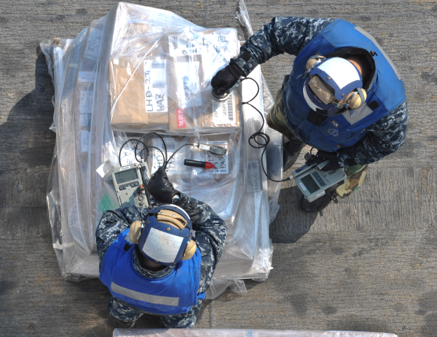 Zwei Männer in blauen Uniformen und Helmen arbeiten an einem großen Drohnenmodell und halten verschiedene Gegenstände, mit einer Plastikabdeckung vor ihnen auf dem Boden.