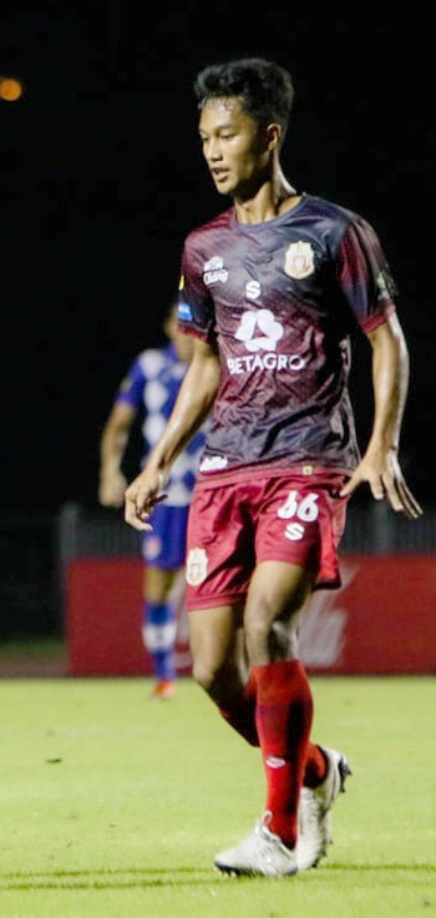 Ein professioneller Fußballspieler in einem maronenfarbenen T-Shirt und roten Shorts, der auf einem Feld spielt, mit einer anderen Person im Hintergrund und einer Tafel mit Text.