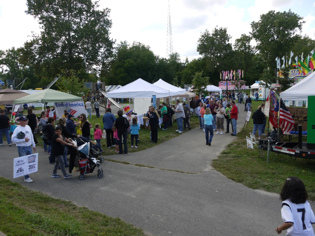 Eine Gruppe von Menschen, Stühle, Fahnen, Baby-Stühle, Zelte und eine Tafel sind auf einer grünen Fläche mit Bäumen im Hintergrund aufgebaut.