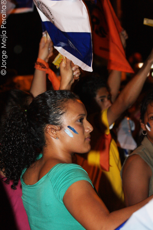 Eine Frau in einem grünen T-Shirt steht auf einer Demonstration und hölt ein weißes Schild, mit vielen anderen Menschen hinter ihr.