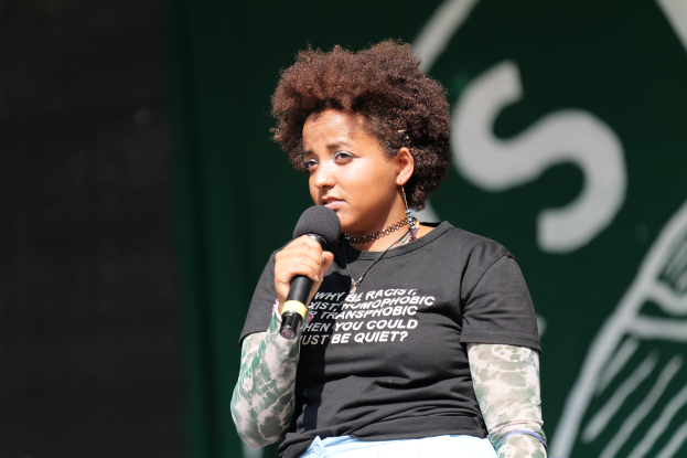 Eine Frau steht selbstbewusst auf einer Bühne, hält ein Mikrofon und trägt ein schwarzes T-Shirt und Jeans, mit einem Pferdeschwanz.