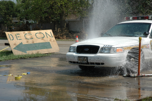Ein Polizeiauto fährt durch eine Pfütze mit verstreuten Gegenständen, darunter eine Tafel mit Text und ein Verkehrskegel, vor einem Hintergrund aus Bäumen, Gebäuden und einem klaren Himmel.