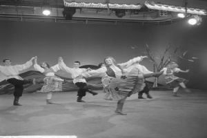 Eine Gruppe von Menschen, die auf einer Bühne tanzen, beleuchtet von Deckenlampen, mit einer Wand im Hintergrund, dargestellt in Schwarz-Weiß.