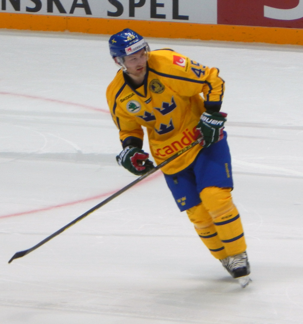 Eishockeyspieler in Bewegung auf dem Eis, trägt Helm, Handschuhe und Schlittschuhe, hält einen Eishockeyschläger und hat eine Tafel mit Text im Hintergrund.