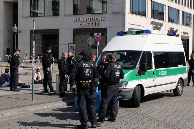 Eine Gruppe von Polizeibeamten steht vor einem Starbucks-Café, rechts daneben parkt ein Van, und links stehen und sitzen einige Menschen. Im Hintergrund ist ein Gebäude mit Fenstern, ein Schild, ein Laternenmast und ein Zaun zu sehen.