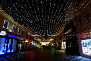 Eine nächtliche Straßenansicht mit Gebäuden zu beiden Seiten, die mit festlichen Weihnachtslichtern geschmückt sind, einige Menschen auf dem Weg und ein dunkler Hintergrund.