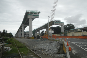 Baustelle mit Brücke im Hintergrund, Straße mit Verkehrskegeln markiert, Schienenstrang links, verstreute Steine und Gras, Bäume und Gebäude säumen die Straße und ein bewölkter Himmel.