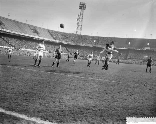 Eine Gruppe von Männern, die auf einem Feld mit einem Stadion, Flutlicht und einem klaren Himmel Fußball spielen.