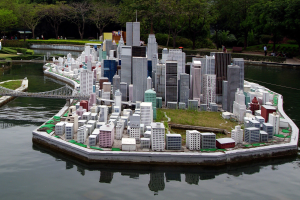 Ein Miniaturmodell einer Stadtlandschaft mit Gebäuden, Grünflächen, einer Brücke und Menschen, auf Wasser gesetzt mit einem Himmelhintergrund.