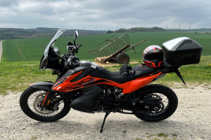 Ein KTM 1290 Super Adventure R Motorrad auf einem Schotterweg geparkt, mit einem Helm und einem Gepäckwagen daneben, umgeben von grünem Gras, hohen Bäumen und Strommästen mit Drähten unter einem bewölktem Himmel.
