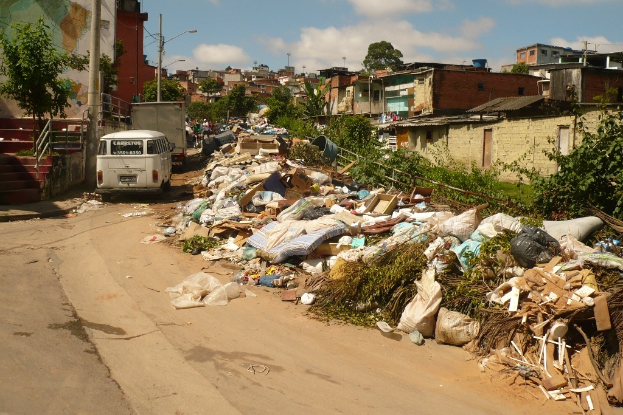Schrott und Pflanzen im Vordergrund, Häuser auf der rechten Seite, Gebäude, Masten, Fahrzeuge und Bäume im Hintergrund unter einem bewölkten Himmel.