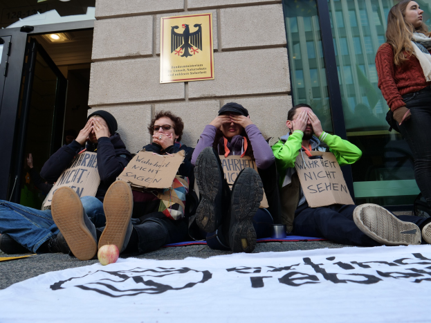 Eine Gruppe von Menschen sitzt vor einem Gebäude auf dem Boden, hält Schilder und hat eine Fahne auf der Straße und eine Frau steht rechts daneben, offenbar bei einer Protestaktion gegen eine Regierungsentscheidung.