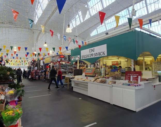Ein belebter Indoor-Markt mit Menschen, die gehen, Blumenbouquets links, Essen auf Tischen rechts, einem Stand mit einem Namensschild und dekorativen Fahnen an der Decke.