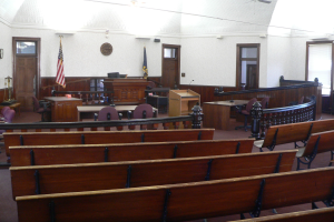 Ein Gerichtssaal mit hölzernen Kirchenbänken, Tischen, Stühlen, einem Rednerpult, Flaggen, Türen, einer Uhr an der Wand und Lampen und Ventilatoren an der Decke.