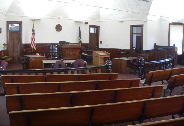 Ein Gerichtssaal mit hölzernen Kirchenbänken, Tischen, Stühlen, einem Rednerpult, Flaggen, Türen, einer Uhr an der Wand und Lampen und Ventilatoren an der Decke.