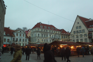 Ein belebter Weihnachtsmarkt in Tallinn, Estland mit Menschen, beleuchteten Ständen, Gebäuden, Bäumen und einem klaren blauen Himmel.