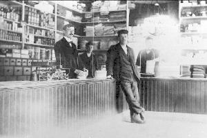 Drei Männer stehen an einem Tresen in einem Geschäft, umgeben von verschiedenen Gegenständen, mit einem Bücherregal und Gegenständen im Hintergrund, die scheinbar an einem Raub beteiligt sind.