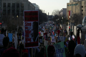 Eine Gruppe von Menschen demonstriert auf einer Straße und hält Plakate, mit Gebäuden, Bäumen und Laternen im Hintergrund unter einem klaren Himmel.