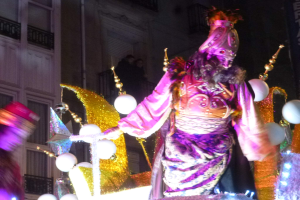 Ein Mann in einem Kostüm steht auf einem dekorierten, beleuchteten Festwagen in einer Stadtparade, im Hintergrund ist ein Gebäude mit Fenstern und Geländern zu sehen.