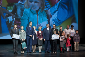 Gruppe von Menschen auf einer Bühne mit Taschen und Urkunden, im Hintergrund ein Bildschirm mit einem Kind und anderen