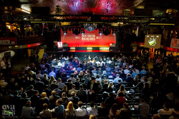 Eine große Menschenmenge sitzt auf Stühlen vor einer Bühne mit Bildschirmen und Plakaten, beleuchtet von Scheinwerfern und umgeben von Absperrungen, mit einem Wasserzeichen in der linken unteren Ecke, und wartet auf ein Online-Marketing-Event für Rockstars.