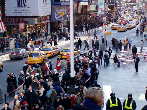 Fahrzeuge fahren auf einer Straße mit Fußgängern auf beiden Seiten, neben Stühlen, Tischen, Laternen, Schildern und Ampeln, mit Gebäuden und Geschäften auf beiden Seiten der Straße, wobei jedes Geschäft ein Schild darüber hat.