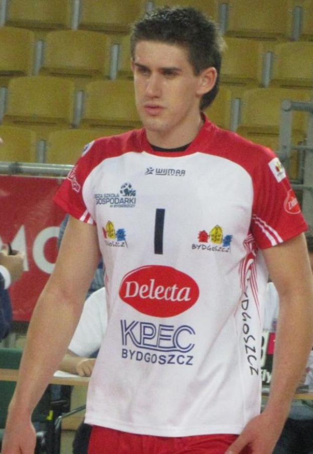 Ein Mann in einer roten und weißen Handballuniform steht auf einem Platz, umgeben von Zuschauern, mit einem roten Banner im Hintergrund, auf dem "Krasnoyarsk vs Bydgoszcz" steht, und sieht entschlossen aus.