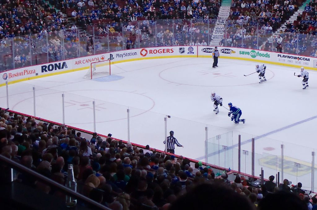Vier Männer beim Eishockey auf einem Eisstadion, mit zwei in der Nähe des Tors und Zauns stehend, Zuschauern auf Stühlen im Hintergrund und eine Treppe sichtbar.