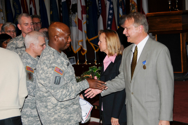 Eine Gruppe von Menschen, die mit zwei Personen Händeschütteln posieren, Flaggen im Hintergrund, ein Rednerpult mit Mikrofon und eine Topfpflanze in der Nähe.